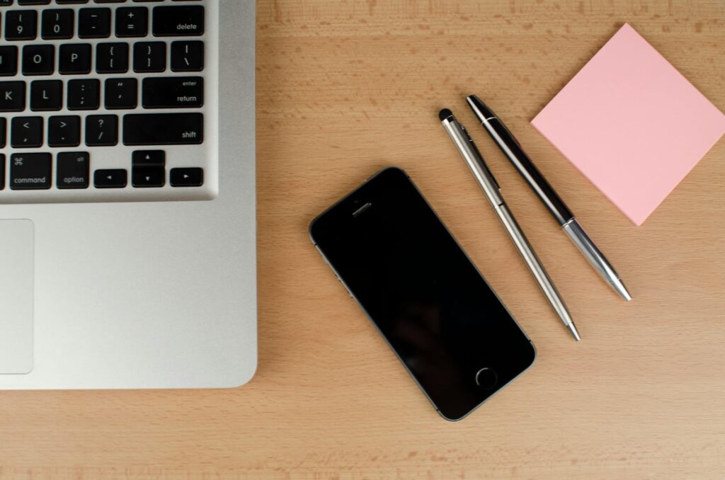 iphone beside, two pens, a sticky note and a laptop